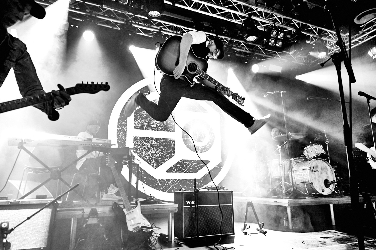 Black and white concert photo: a guitarist leaps high across a smoky stage under the lights, with the band playing behind him.