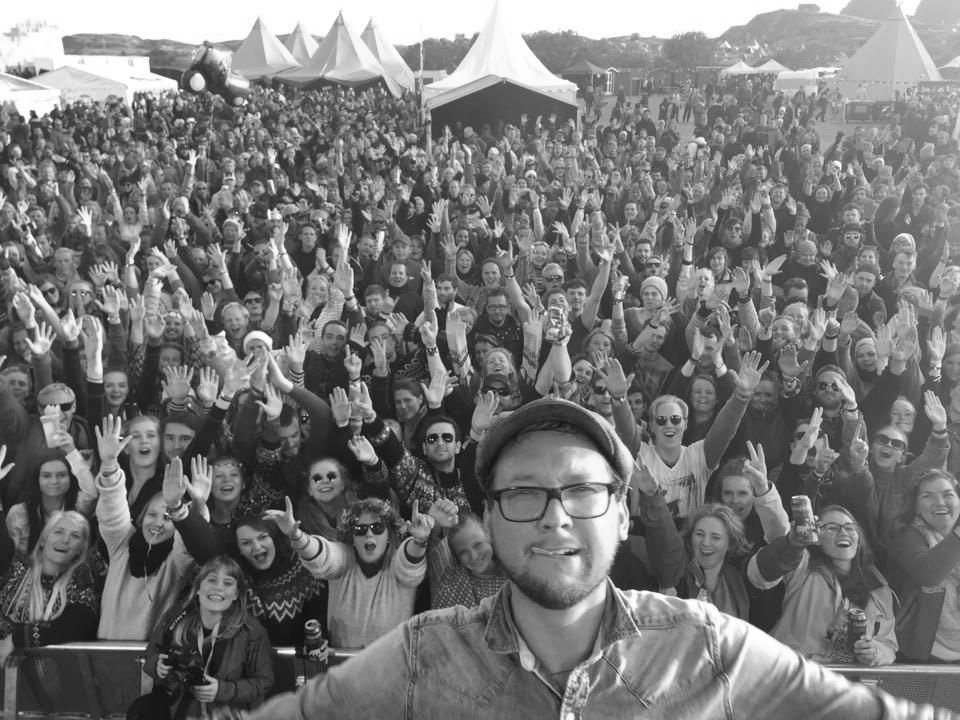 Dan Hesketh taking a selfie from a festival stage, with a large crowd behind him raising their hands.